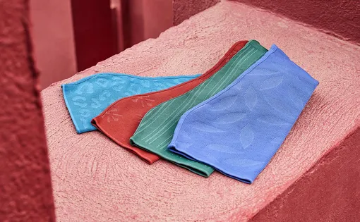 Vier flach ausgebreitete Kompressionsstrümpfe in den Farben Hellblau, Dunkelrot, Smaragdgrün und Königsblau liegen gefächert auf einer rauen, rosafarbenen Steinfläche. Jeder Strumpf weist ein individuelles eingestricktes Muster auf.