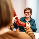 Die Beraterin überreicht der Kundin einen Sockenfuß mit einem rostroten, gemusterten Kompressionsstrumpf von Medi
