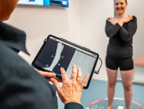 Eine Beraterin bedient im Vordergrund ein Tablet, auf dem das digitale 3D-Modell eines Beines mit Messpunkten zu sehen ist. Im Hintergrund steht die lächelnde Kundin barfuß auf der Vermessungsmatte, während ihre Maße berührungslos erfasst werden.