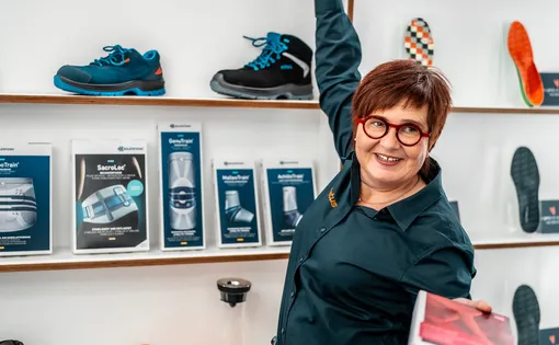 Eine lächelnde Limotus-Beraterin steht vor einem Regal und streckt eine Hand nach einer Verpackung aus, während sie in der anderen Hand eine Schachtel hält. Auf den Regalen sind verschiedene Produkte wie Schuhe und Bandagen zu sehen.