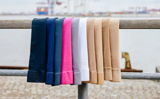 Eine Auswahl an Limotus Kompressionsstrümpfen in verschiedenen Farben, die nebeneinander über einem Holzgeländer hängen. Zu sehen sind Töne wie Dunkelblau, Marine, Anthrazit, Pink, Grau, Weiß sowie verschiedene Beigetöne. Im Hintergrund iein Hafenbecken.