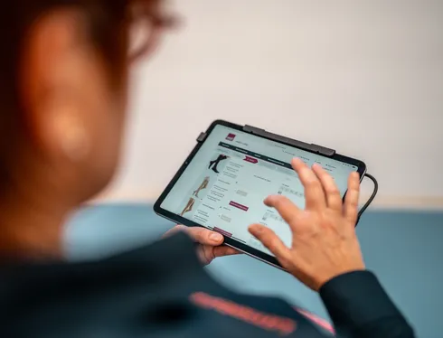 Nahaufnahme über die Schulter einer Mitarbeiterin, die ein Tablet bedient. Auf dem Bildschirm ist ein Online-Konfigurator für Kompressionsstrümpfe mit verschiedenen Produktbildern und Auswahloptionen zu sehen. Der Fokus liegt auf der Hand am Tablet.